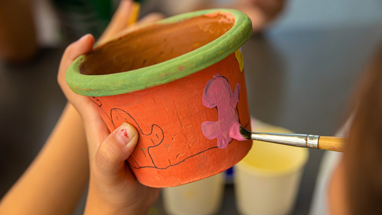 Close-up of a child's hands painting a small clay flower pot with pink paint using a brush.
