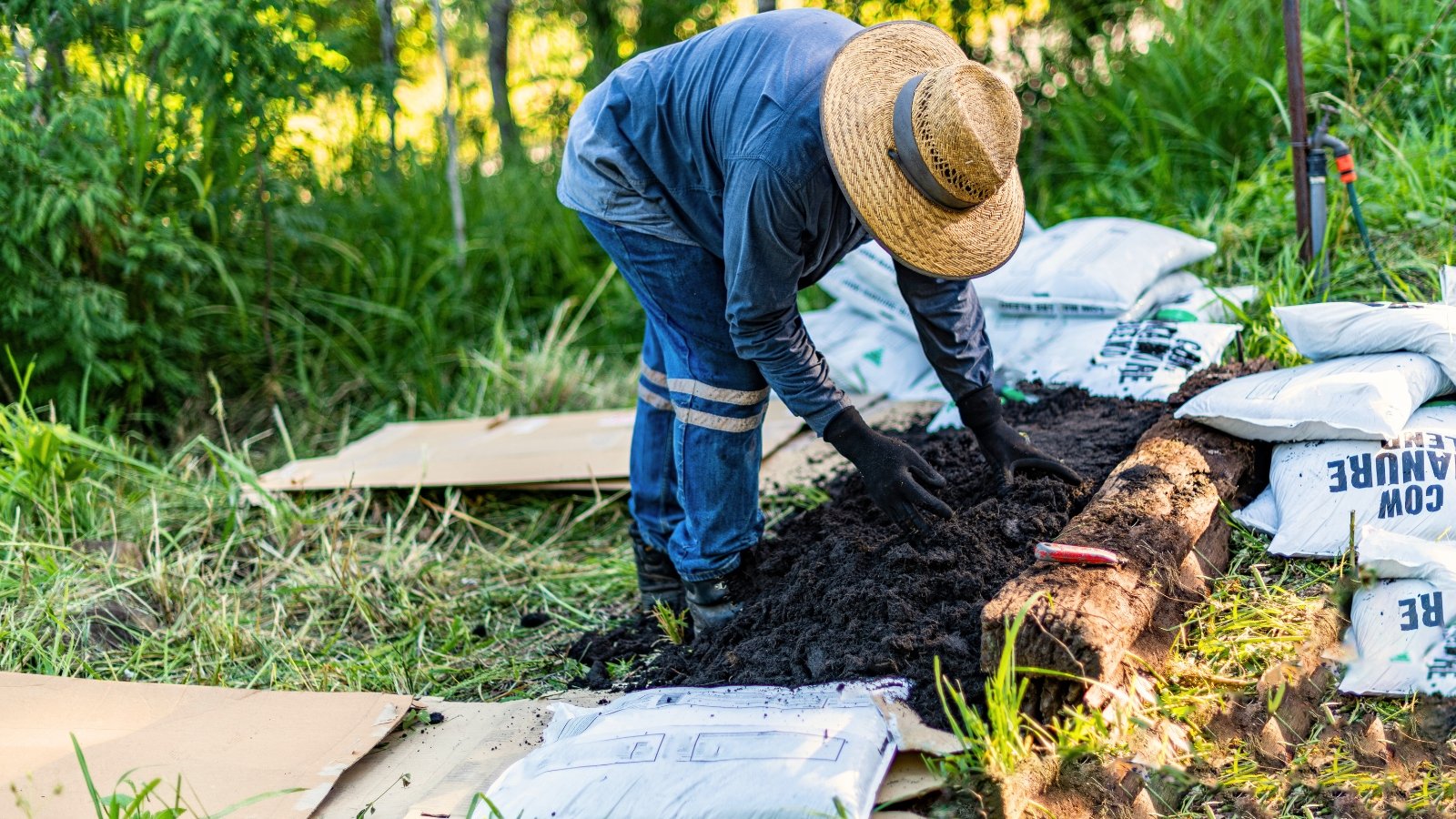 A gardener in black gloves and a wide-brimmed wicker hat spreads fresh black compost on a bed in a garden.