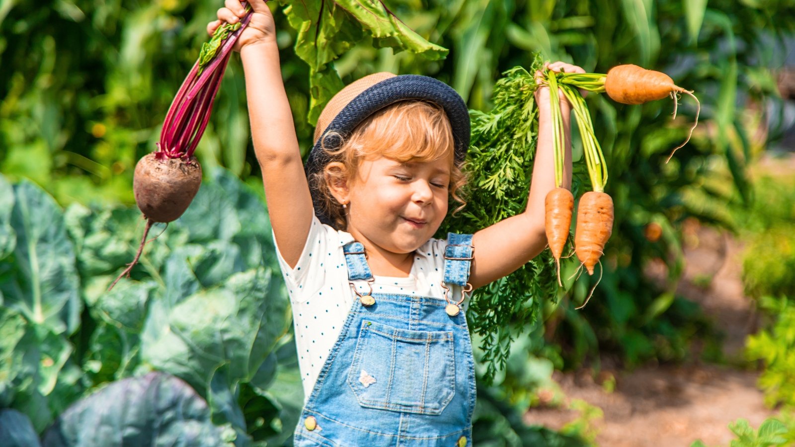 A girl with golden hair, wearing a hat and denim dress, holds freshly picked carrots and beets in a sunny garden.