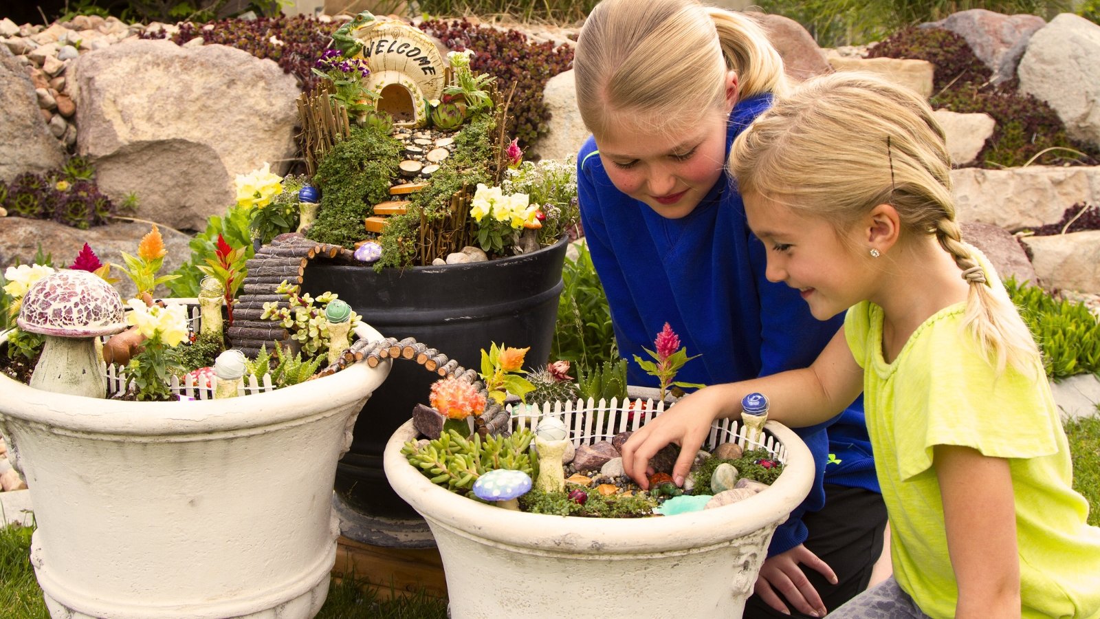 Close up of two blonde girls creating small fairy gardens in white and black large containers.