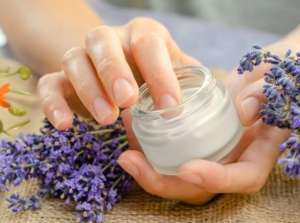 Close-up of female hands holding a small jar of homemade cream over a table with fresh blooming sprigs of lavender and calendula.