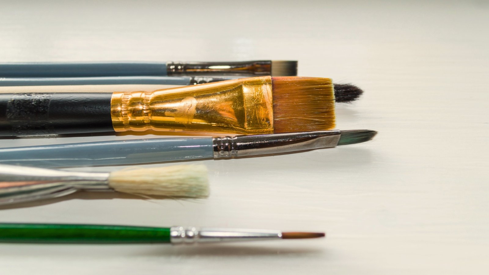 Paint brushes with long, slender handles and bristles of varying lengths and shapes, arranged in fan and flat configurations at the tips, rest on a white wooden table.