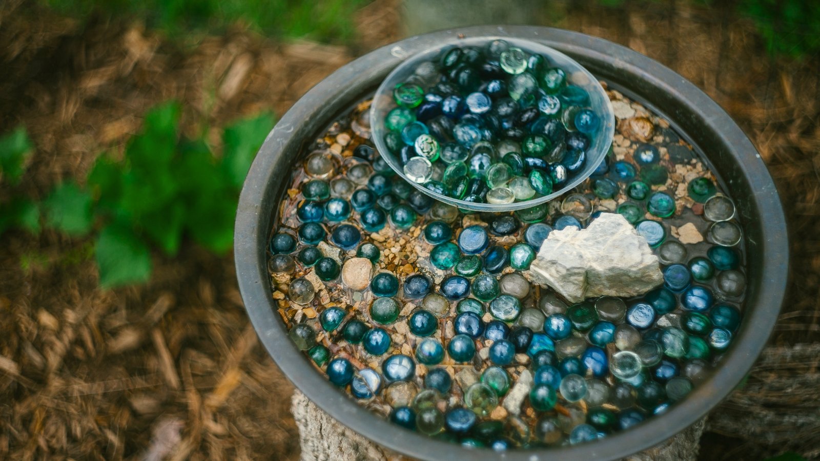 The Pollinator Watering Station features a shallow, decorative basin filled with water and surrounded by stones and pebbles, designed to provide hydration for bees and other pollinators.