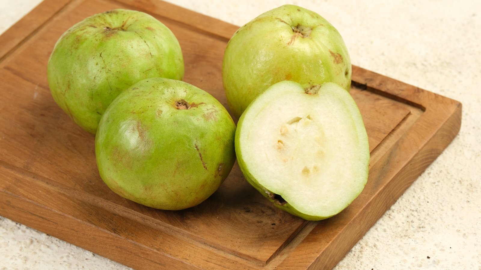 Close-up of three whole ripe guavas and a half-cut fruit, showing crisp light green skin and white, juicy flesh.
